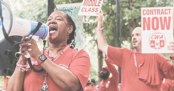 A person in a red shirt holding a bullhorn with people in red shirts holding signs that say Contract Now.